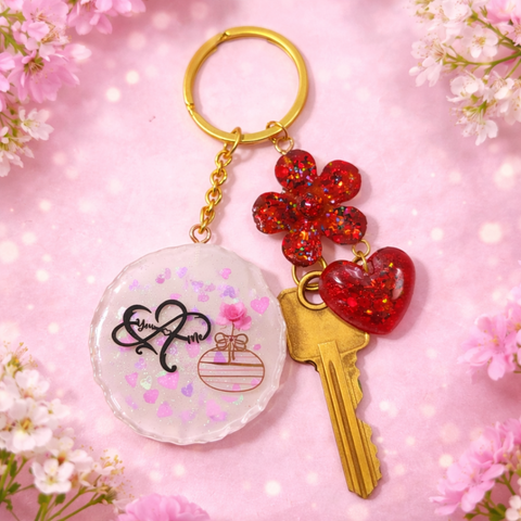 A personalized white resin keychain with a "You & Me" design and pink hearts, shown with a gold key attached alongside a red glittery flower and heart charm against a floral pink background.