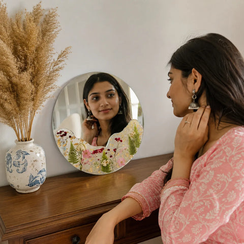 Woman adjusting earrings in front of a round mirror with floral design, next to a vase with dried plants.