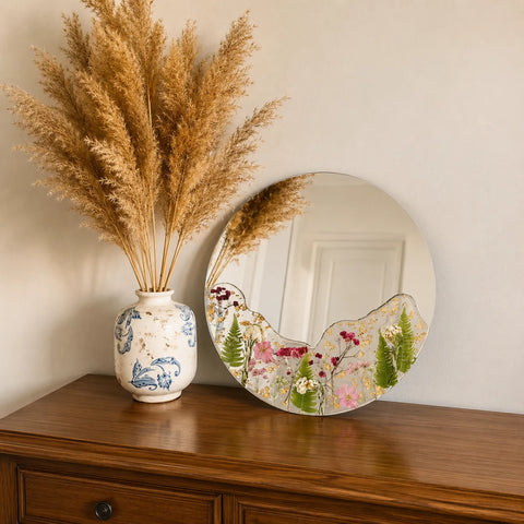 Decorative vase with dried pampas grass and floral-patterned mirror on a wooden surface.